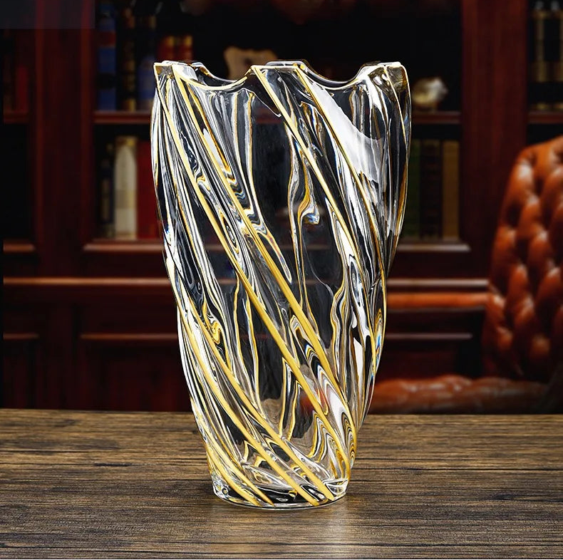 A clear glass vase with a wavy, organic gold swirl pattern, displayed on a dark wooden table with a blurred background of a bookshelf and leather chair.
